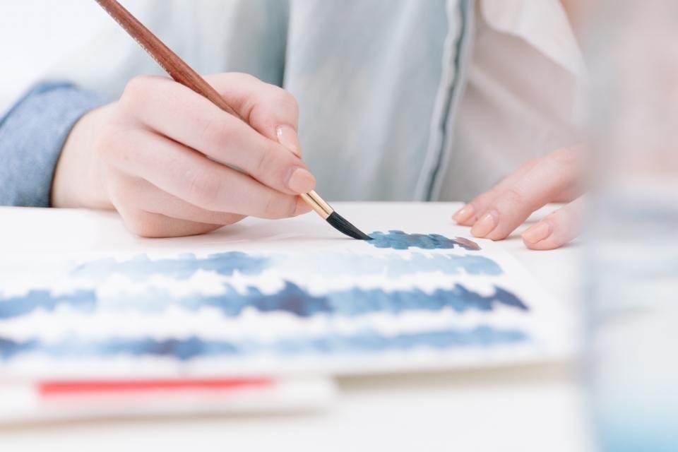 Mão segurando um pincel e pintando faixas de aquarela azul sobre papel branco